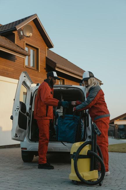 Two movers from Storage Wandsworth are engaged in a home relocation process, loading a large, dark green upholstered piece of furniture into the back of a white van parked on the street. The van's rea