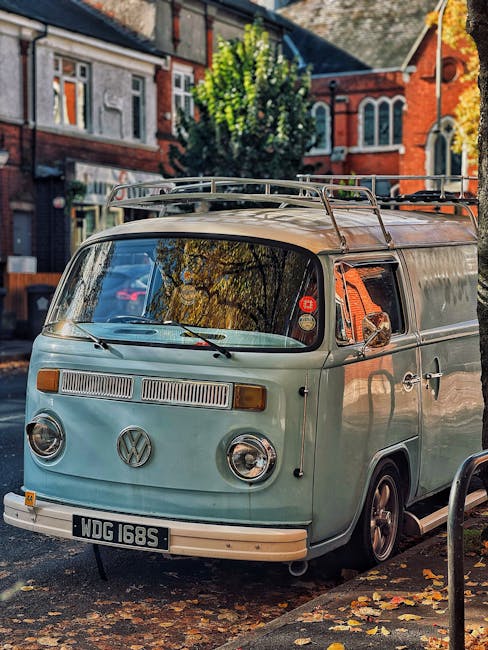 A vintage light green van with tinted windows and black tires is parked on a city street in front of a white building, surrounded by tall leafy trees with rich green foliage. The van is positioned par