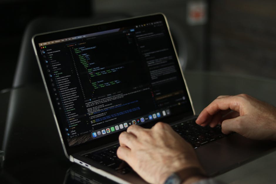 Close-up of a person's hands typing on a laptop with a dark screen displaying lines of code and text, placed on a black table in a well-lit indoor setting. The individual is engaged in a computer-base