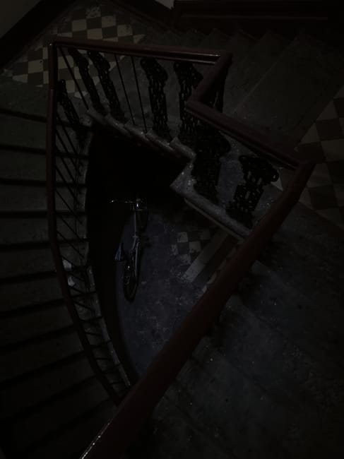 A view from above of a narrow staircase inside a Victorian home, featuring ornate black wrought iron railings with decorative scrollwork and a polished wooden handrail. The staircase is covered by a r