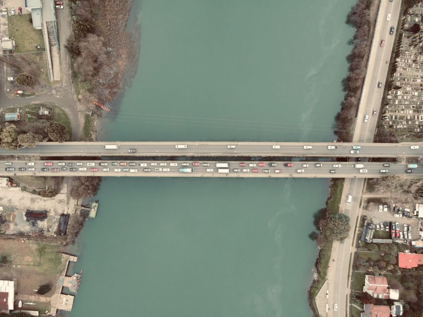 An aerial view showing a bridge crossing over a wide river, with multiple lanes of traffic including cars, vans, and trucks moving in both directions. The bridge is constructed with concrete and road