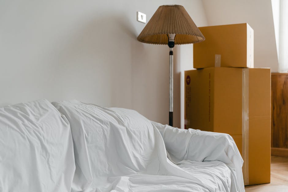A man wearing a black cap and a white polo shirt is standing indoors next to a large stack of uniformly-sized cardboard boxes, some of which are stacked on top of each other against a plain wall. The 