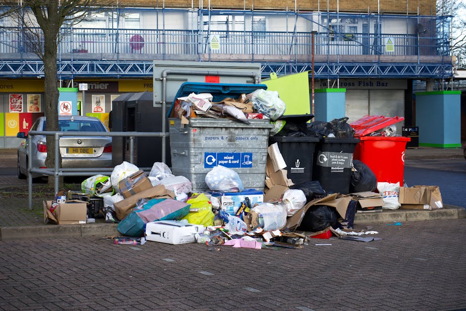 A street scene showing multiple overflowing waste and recycling bins, with black, black, and red plastic bags, cardboard boxes, and paper packaging scattered around the bins and on the pavement. The r
